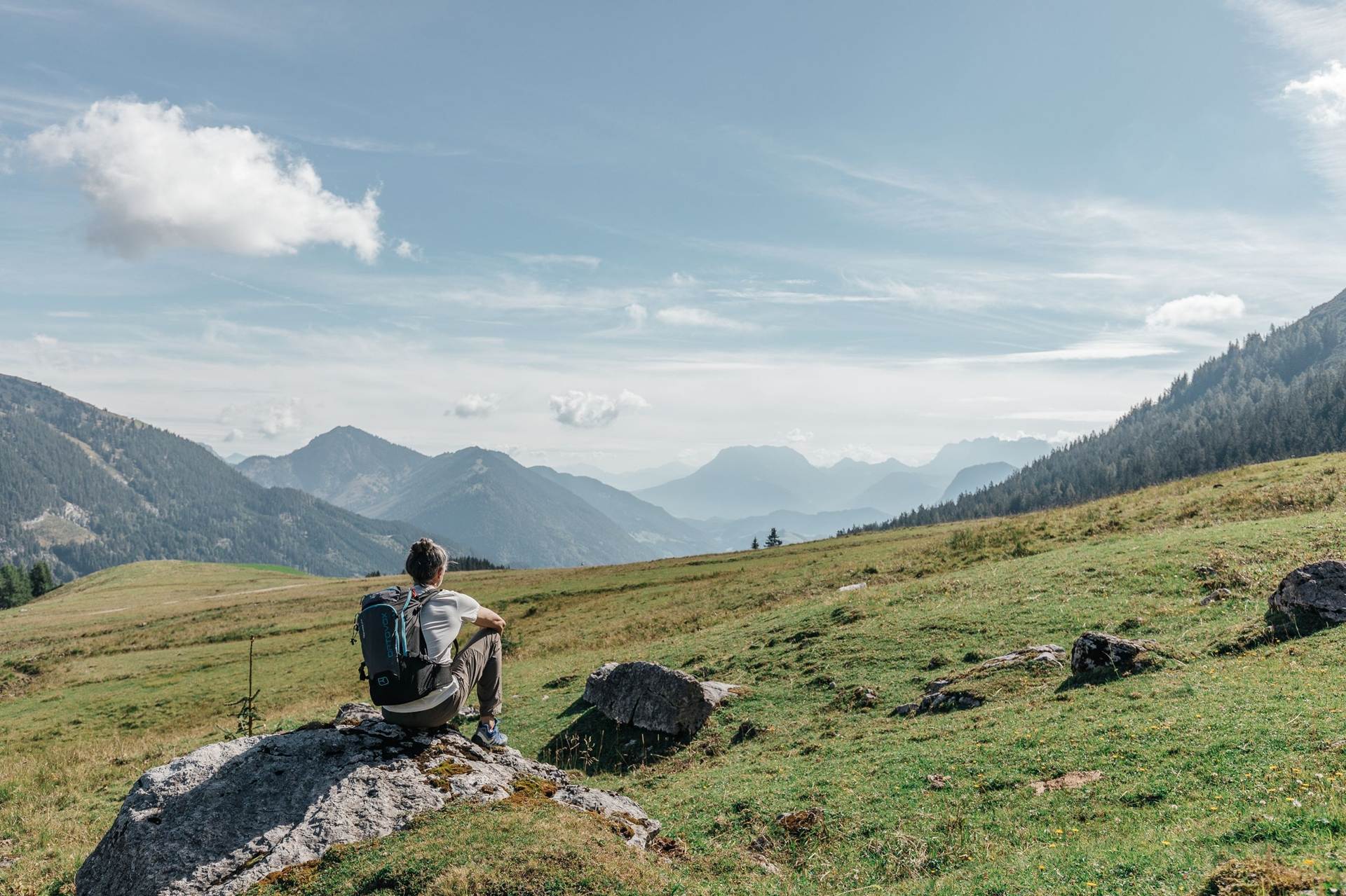 Sommerurlaub in den Bergen verbringen Ein Wanderer sitzt auf einem Stein vor einer weiten Wiese umgeben von Bergen.
