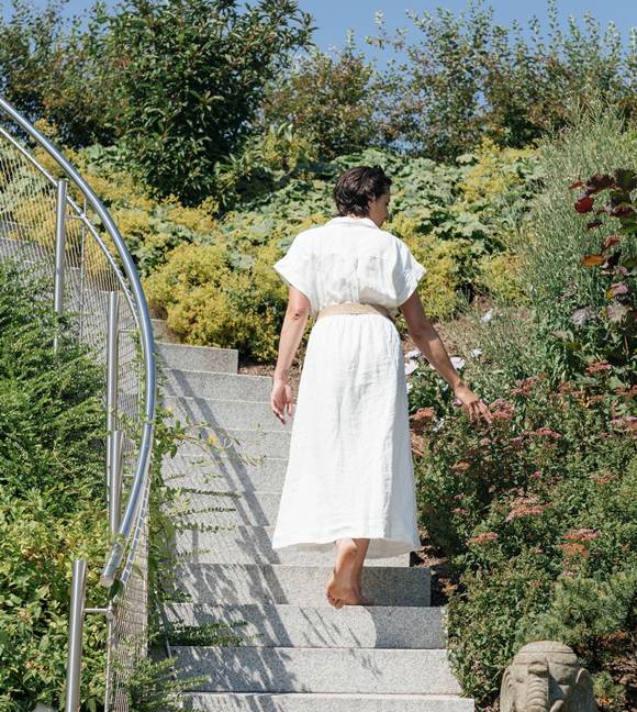 A woman in a white dress is walking up some steps in the garden and reaching out towards the bushes beside her.