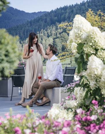 Ein Mann sitzt auf einer Bank, eine Frau steht neben ihm, beide halten ein Glas Wein und sehen sich an. Sie sind umgeben von Blumen.