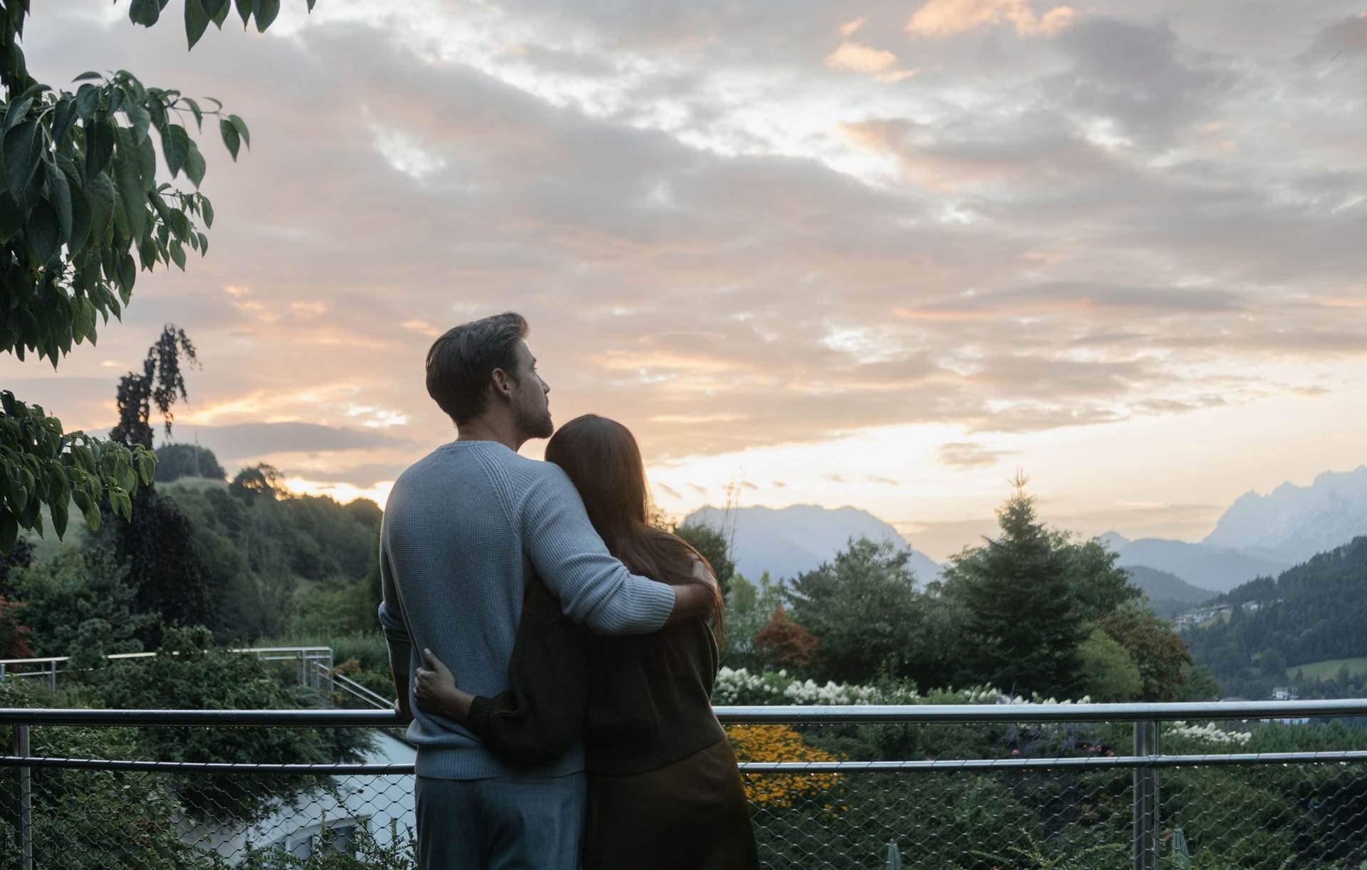 Im Juffing Hotel & Spa erleben Sie unvergessliche Momente harmonischer Zweisamkeit Ein Paar steht Arm in Arm am Balkon und schaut in die umliegende Landschaft.