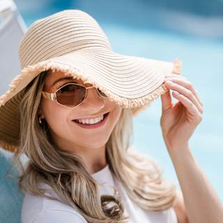 Eine lachende Frau mit Sonnenhut und Sonnenbrille, hinter ihr der Pool.